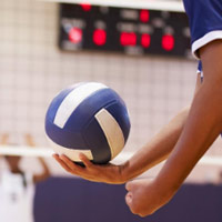 Volleyball in einer Turnhalle. Im Vordergrund ist der Körper einer Person mit einem Volleyball in der Hand. Im Hintergrund ein Volleyballnetz und hinter dem Netz drei andere Spieler verschwommen. An der Wand ist verschwommen eine Anzeigetafel zu sehen.