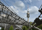 Auf diesem Bild ist das Dach im Olympiapark und der Olympiaturm zu sehen. Im Hintergrund sind Bäume und ein Himmel mit Wolken zu sehen. 