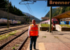 Hier steht Herr Staatsminister Christian Bernreiter an einem Gleis am Brenner. Der Himmel ist blau. Im Hintergrund sind Gleise und ein roter Zug zu sehen. 