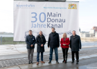 Bayernhafen Geschäftsführer Joachim Zimmermann, Umweltminister Thorsten Glauber, Ministerpräsident Dr. Markus Söder, Europaministerin Melanie Huml und Verkehrsminister Christian Bernreiter stehen vor einem großen Plakat mit der Aufschrift "30 Jahre Main-Donau-Kanal". Im Hintergrund ist der Bamberger Hafen zu sehen.