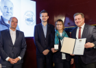 Verkehrsminister Christian Bernreiter überreicht eine Urkunde an eine Frau und einen Mann. Daneben steht der Laudator. Im Hintergrund ist das Foto eines Mannes auf der Leinwand zu sehen.