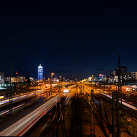 Blick von oben auf die Gleise zum Münchner Hauptbahnhof bei Nacht. Es sind beleuchtete Häuser und Lichter sowie verschwommen fahrende Züge zu erkennen.