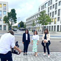 Michaela Ausfelder, die verantwortliche Architektin des Mehrgenerationenwohnens an der Katharina-Mair-Straße, Antonia Ivanković, Referentin für Wohnraumförderung an der Regierung von Obernbayern und Barbara Schelle, Stadtbaumeisterin der Stadt Freising stehen vor einem Wohngebäude in Freising und werden dabei von einer Kamera gefilmt