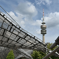 Auf diesem Bild ist das Dach im Olympiapark und der Olympiaturm zu sehen. Im Hintergrund sind Bäume und ein Himmel mit Wolken zu sehen. 