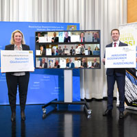 Gruppenbild der 15 neu ausgezeichneten fahrradfreundlichen Kommunen  (am Bildschirm) mit Bayerns Verkehrsministerin Kerstin Schreyer und AGFK-Vorsitzendem Matthias Dießl, die jeweils mit einem Gratulationsschild links und rechts vom Bildschirm stehen 