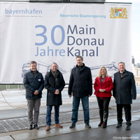 Bayernhafen Geschäftsführer Joachim Zimmermann, Umweltminister Thorsten Glauber, Ministerpräsident Dr. Markus Söder, Europaministerin Melanie Huml und Verkehrsminister Christian Bernreiter stehen vor einem großen Plakat mit der Aufschrift "30 Jahre Main-Donau-Kanal". Im Hintergrund ist der Bamberger Hafen zu sehen.