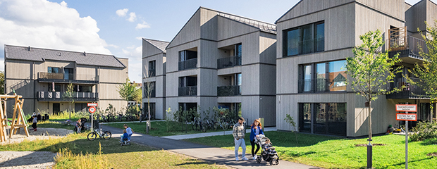 Das Bild zeigt die Bewohnerschaft in mitten einer modernen und autofreien Wohnanlage mit gemeinschaftlichen Grünflächen und einem Spielplatz.